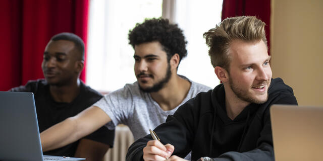 Three students at a table in a classroom with laptops in front of them.