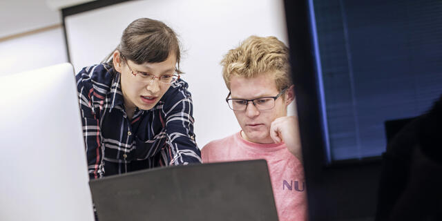 A professor leans over a student's shoulder while they look at a computer screen together.