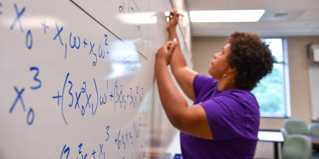 A Black, female student writes equations on a whiteboard.