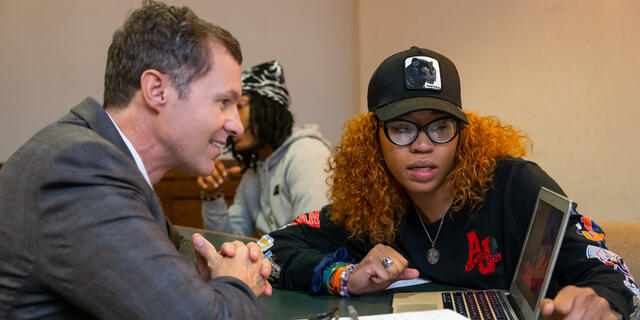 A Black, female student speaks to a white man while they look at a laptop screen.