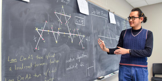 A male student gestures to a blackboard where a math equation is written.
