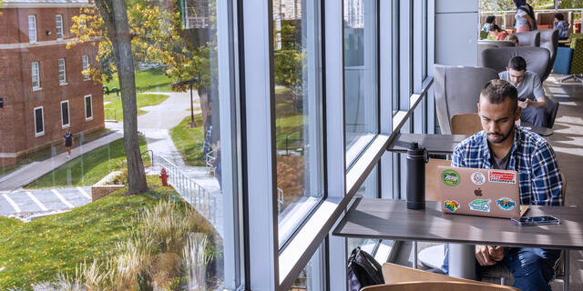 A student studies on a laptop a large window in the Roj Student Center.