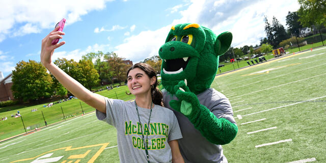 A female student in a McDaniel College t-shirt poses with the Green Terror mascot while taking a photo on her phone.
