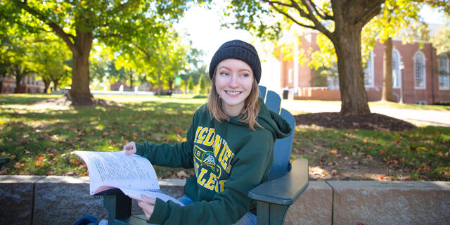 A female student sits on a lawn chair on campus while holding a book and smiling at the camera.