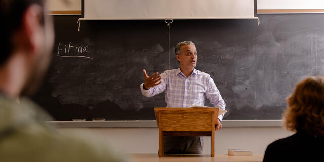 A male faculty member stands at the front of a classroom behind a podium while gesturing with his hand.