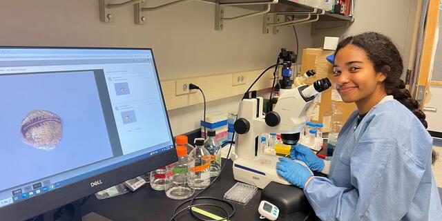 A student in a blue lab coat smiles at the camera while sitting in front of a microscope. An image of a zebrafish embryo from under the microscope appears on a computer screen next to her.