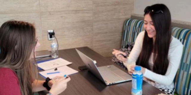 Two students sit at a booth table together while sampling food and looking at laptops and notebooks in front of them.