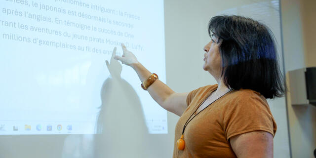 A McDaniel French professor in an orange shirt points at a projector screen with the French language on it.