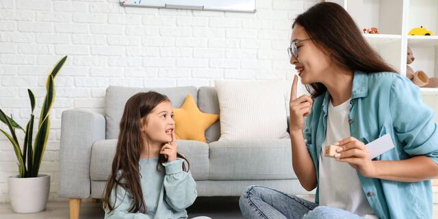 A woman and child sit crossed legged on the floor in front of a couch, each touching a finger to their mouths.