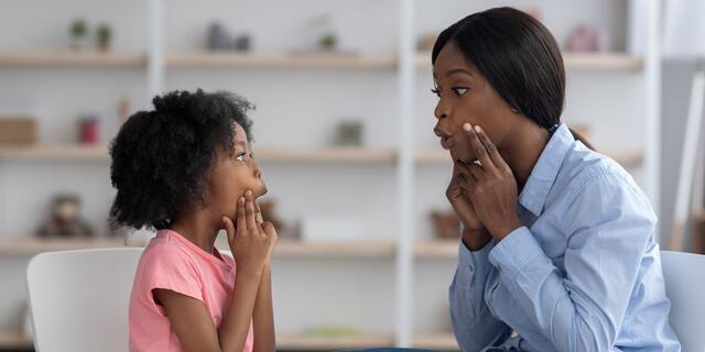 A young woman sits next to a small child at a table. They both hold their hands up to their face while shaping a sound with their mouths.