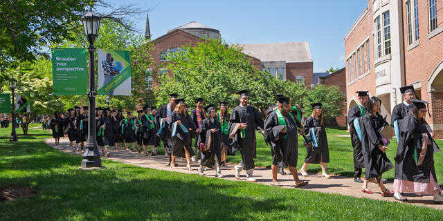 A line of graduate students in regalia walk down a path across the McDaniel campus in the spring.