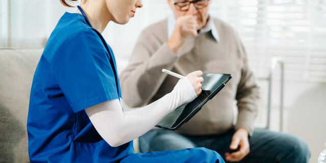 A young woman in blue scrubs writes on a clipboard as a senior man seated next to her speaks.