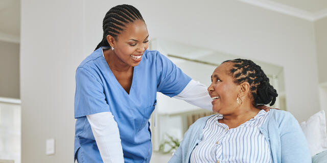A young African American woman in scrubs leans over a senior African American woman patient while they both smile.