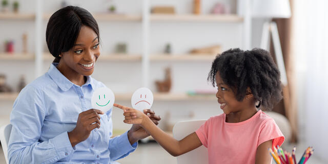 A school counselor holds up a paper smiling face and a paper frowning face, and a young student points at the smiling face.