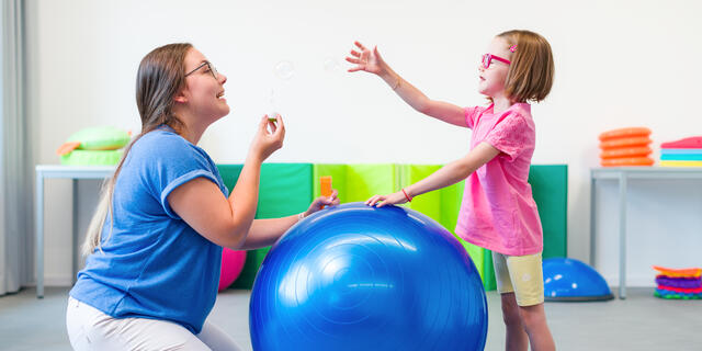 Occupational therapist kneels on the ground with a large blue ball and a young girl in pink