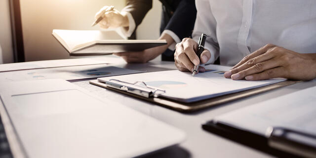 Two people sit at a desk covered in paperwork. One holds a pen and the other holds a book.
