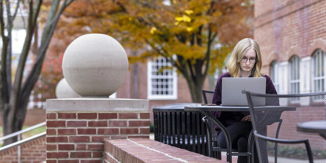 A female student sits at an outdoor table and types on a laptop.