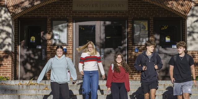 group of students walk down the steps Fall leaves and trees in the background in front of a building with a sign reading "Hoover Library". 