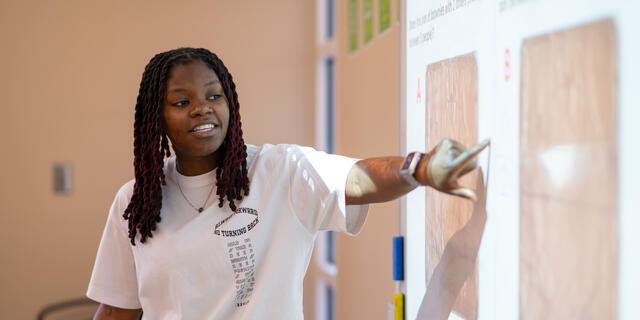 A young student teacher in a white shirt points to a projection on a wall.