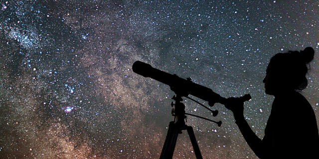 A silhouette of someone pointing a telescope at a sky full of stars.