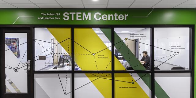 Entrance to The Robert '92 and Heather Pick STEM Center, with green signage above glass windows decorated in yellow and green diagonal stripes and geometric line graphics reading "Proof that problem-solving is a team sport," "It's break, then make time," "Pi: More than just dessert," "Sharpen your skills—no lab goggles required," and "Turning 'Huh?' into 'Aha!' moments." Through the windows, a person is visible seated at a desk near a whiteboard and computer workstations.