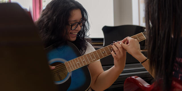 A student with dark hair and glasses plays a guitar with help from an instructor.